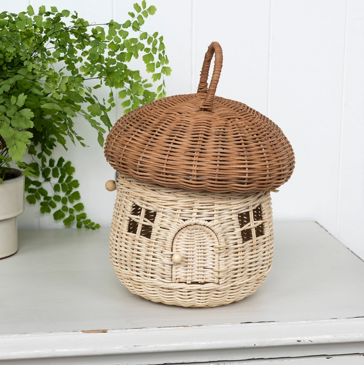 Rex London Wonders of Nature rattan mushroom house decorative basket — lifestyle shot on a white wooden surface beside a maidenhair fern in a white pot; showing the full mushroom house with honey-brown domed cap, pale cream woven body with lattice windows and arched doorway, and looped rattan carry handle