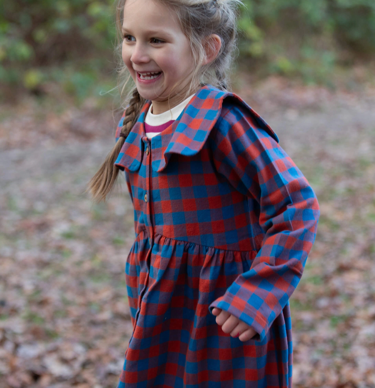 Little Green Rascals burnt ochre and teal blue check sailor dress — lifestyle shot of a young girl laughing and running outdoors in autumn woodland, wearing the dress with a cream top layered underneath; showing the full gathered skirt in motion and the wide flat sailor collar