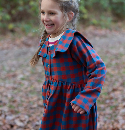 Little Green Rascals burnt ochre and teal blue check sailor dress — lifestyle shot of a young girl laughing and running outdoors in autumn woodland, wearing the dress with a cream top layered underneath; showing the full gathered skirt in motion and the wide flat sailor collar