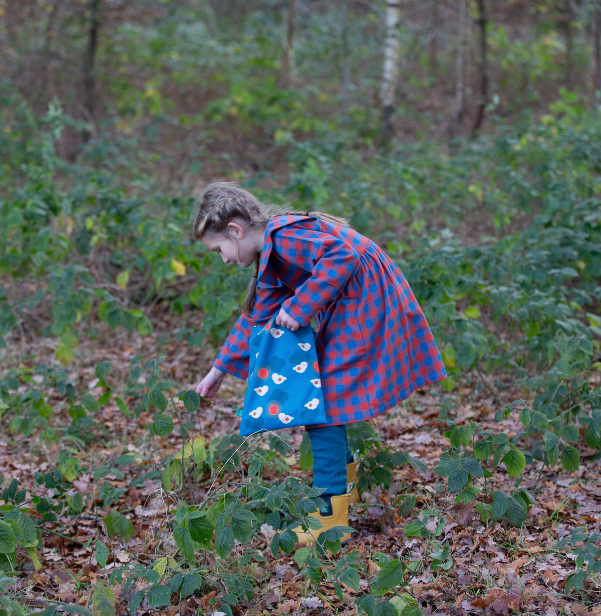 Little Green Rascals burnt ochre and teal blue check sailor dress — lifestyle shot of a young girl exploring in autumn woodland wearing the dress with yellow wellies and carrying a blue tote bag; showing the full length and swishy gathered skirt