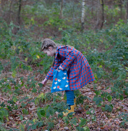 Little Green Rascals burnt ochre and teal blue check sailor dress — lifestyle shot of a young girl exploring in autumn woodland wearing the dress with yellow wellies and carrying a blue tote bag; showing the full length and swishy gathered skirt