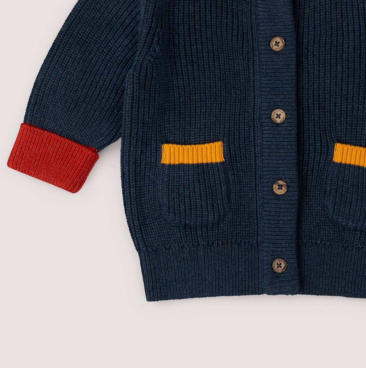 Little Green Rascals Midnight Snuggly Knitted Cardigan — close-up of the lower body showing the two front patch pockets with mustard yellow ribbed trim at the top, branded coconut shell buttons down the front placket, red ribbed turned-up cuff, and ribbed hem