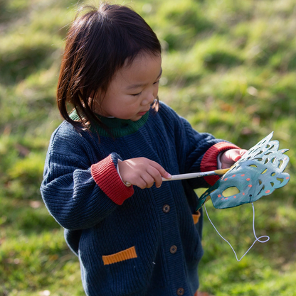 Little Green Rascals Midnight Snuggly Knitted Cardigan — lifestyle shot of a young toddler girl outdoors in a sunny garden wearing the midnight navy cardigan with forest green neckline, red cuffs and mustard pocket trim, holding a decorative butterfly on a stick