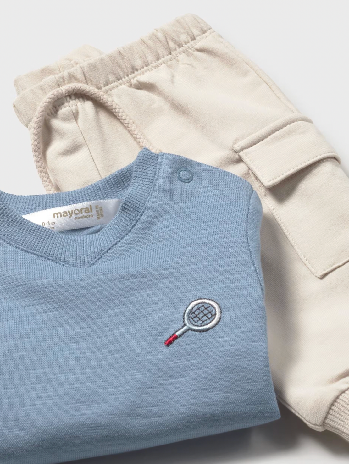 Mayoral boys Tennis Blues 2-piece set — close-up detail showing the cornflower blue pullover folded to reveal the embroidered tennis racket motif in navy and red on the left chest, Mayoral label at the neckline and a snap button shoulder fastening; sand beige cargo joggers with large patch cargo pocket and ribbed ankle cuff visible behind