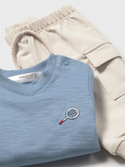 Mayoral boys Tennis Blues 2-piece set — close-up detail showing the cornflower blue pullover folded to reveal the embroidered tennis racket motif in navy and red on the left chest, Mayoral label at the neckline and a snap button shoulder fastening; sand beige cargo joggers with large patch cargo pocket and ribbed ankle cuff visible behind