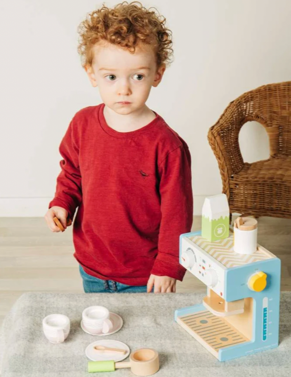 Bigjigs wooden espresso coffee maker — lifestyle shot of a young curly-haired boy in a red jumper standing at a grey linen-covered surface with the wooden coffee machine set up beside him; pale blue machine panels, yellow toggle switch, green milk carton and white sugar pot visible on and beside the machine; two blush pink cups on saucers, a pink saucer, the natural wood milk jug and green portafilter handle arranged on the surface in front; wicker chair visible in the background