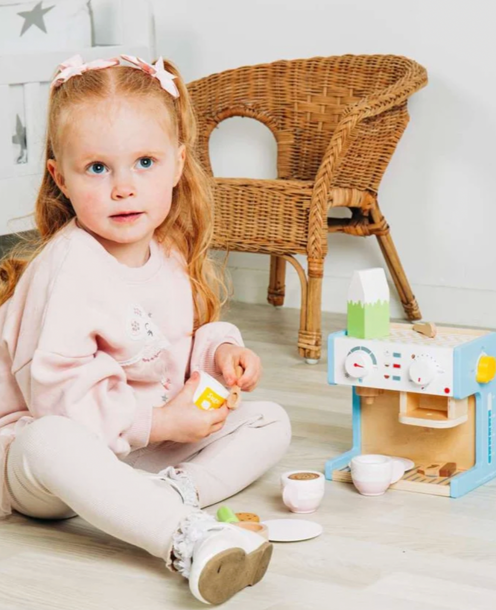 Bigjigs wooden espresso coffee maker — lifestyle shot of a young red-haired girl in a pink sweatshirt sitting cross-legged on a light wood floor, holding the yellow sugar pot and playing barista; the wooden coffee machine with pale blue panels, green milk carton on top and yellow toggle switch is set up beside her; blush pink cups, saucers and the green portafilter handle are arranged on the floor in front; wicker chair and star-print wall visible in the background
