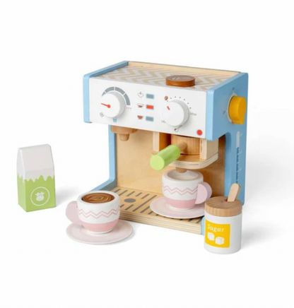 Bigjigs wooden espresso coffee maker play set — three-quarter view on white background showing the full set from a slightly elevated angle; natural birch wood machine body with pale blue side panels, white front panel with dials and indicator lights, natural wood circular lid on top, yellow toggle switch on right side, green portafilter handle in the group head; accessories displayed: green and white milk carton (left), two blush pink chevron-pattern cups on pink saucers (front and on drip tray), white suga