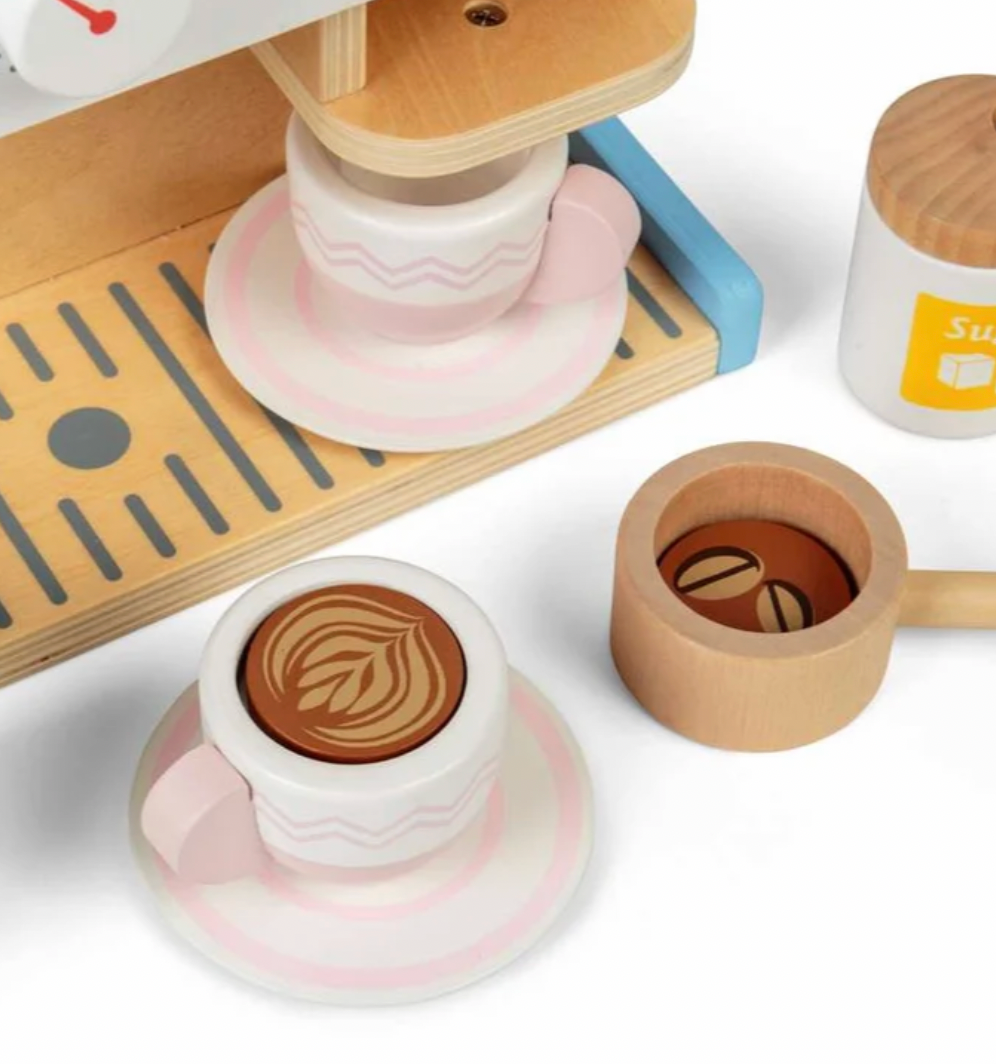  Bigjigs wooden espresso coffee maker — close-up detail showing a blush pink chevron-pattern cup on a pink saucer with a beautifully printed latte art leaf swirl in brown on white inside the cup (foreground left); a natural tan wood handled milk jug/pitcher with a coffee bean motif printed inside (foreground right); natural wood slatted drip tray with a second blush pink cup on saucer and white sugar pot with natural wood lid visible in the background