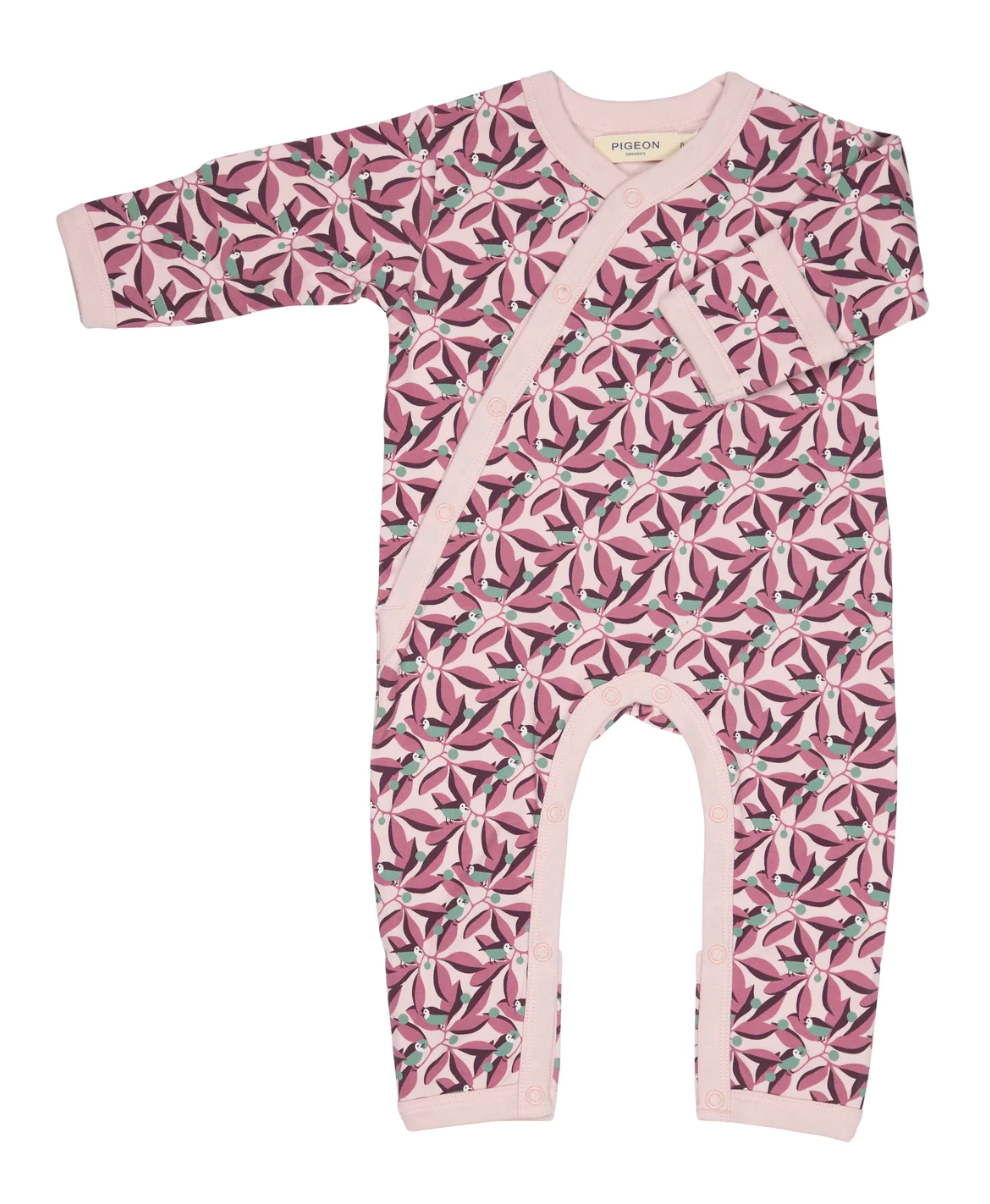 Front flat — pale blush ground; dense plum and mauve tropical foliage; teal songbirds and berry clusters throughout; kimono wrap; fold-over cuff; leg poppers; blush ribbed trims; Pigeon Organics label.
