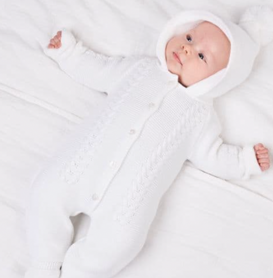 Newborn baby lifestyle — lying on white textured blanket; looking upward; hood framing face; cable knit front; bootees; serene all-white aesthetic.