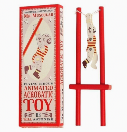 Left: tall red and cream vintage circus gift box reading "Flying Circus / Animated Acrobatic Toy / He Will Astonish"; right: assembled toy with figure hanging inverted