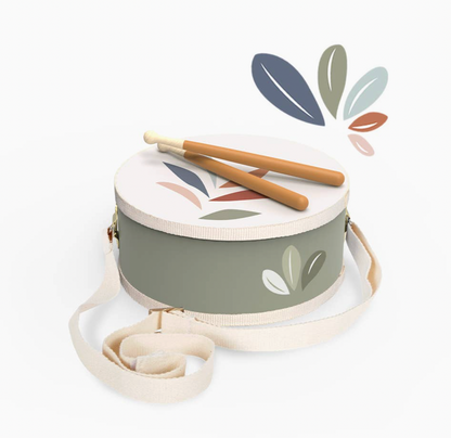 Angled overhead product shot — sage green drum, white botanical leaf head, two drumsticks crossed on top, cream strap trailing; decorative leaf graphic top right