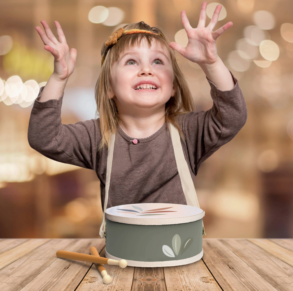 Lifestyle — girl wearing drum on strap, arms thrown up with pure joy; drumsticks on table; warm bokeh background
