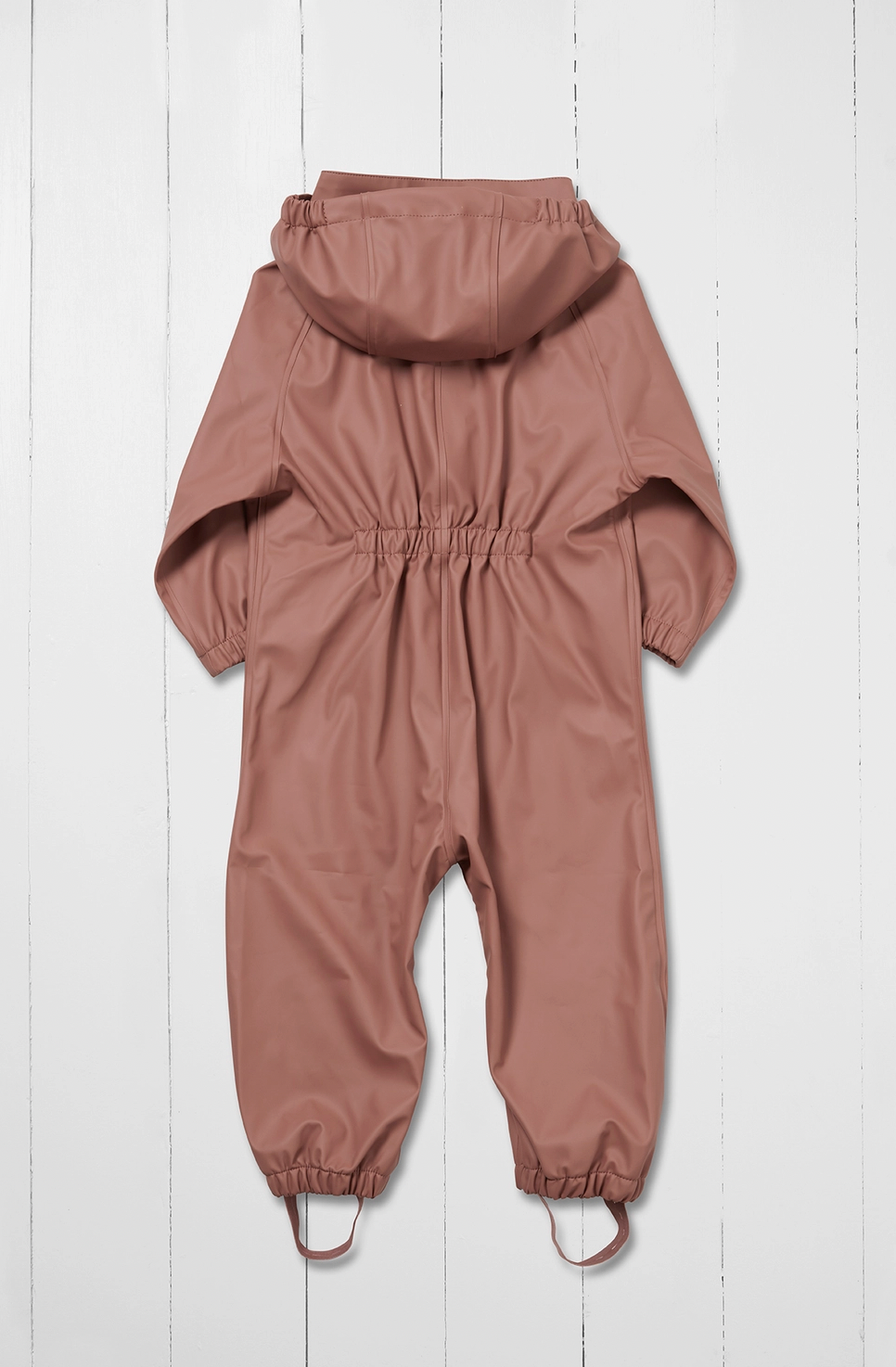 Grass & Air kids waterproof puddle suit in muted dusty rose shown as a rear flat lay against a white wooden plank background; full one-piece all-in-one suit viewed from the back in warm clay pink; attached hood visible at the top showing the rear panel; prominent elasticated gathered back waist panel creating shape across the centre back; long sleeves with elasticated gathered cuffs visible at the sides; full legs with elasticated gathered ankle cuffs at the bottom; integrated boot stirrups in matching rose