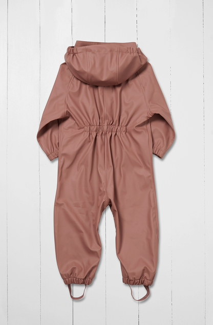 Grass & Air kids waterproof puddle suit in muted dusty rose shown as a rear flat lay against a white wooden plank background; full one-piece all-in-one suit viewed from the back in warm clay pink; attached hood visible at the top showing the rear panel; prominent elasticated gathered back waist panel creating shape across the centre back; long sleeves with elasticated gathered cuffs visible at the sides; full legs with elasticated gathered ankle cuffs at the bottom; integrated boot stirrups in matching rose