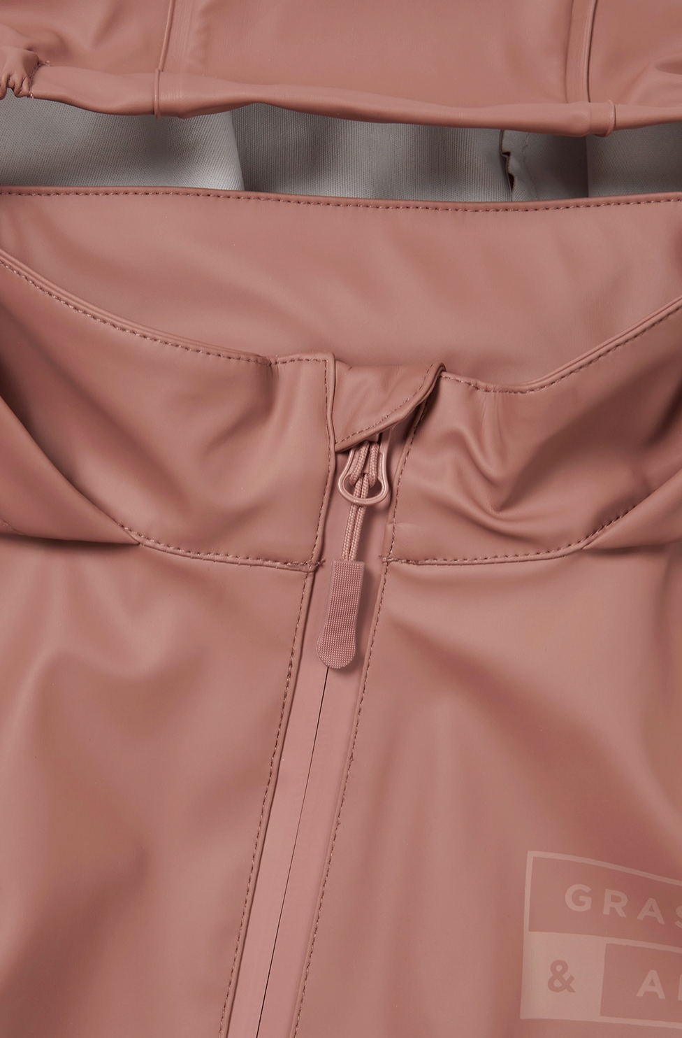 Close-up detail of the back waist panel of the Grass & Air Rose puddle suit; wide horizontal elasticated back waist panel gathered and stitched across the back of the suit creating shape and allowing a generous comfortable fit over clothes and layers; rose waterproof fabric gathered either side of the central elasticated band; taped seam stitching visible; tone-on-tone Grass & Air logo label printed in matching rose visible in the bottom right corner; zip guard flap partially visible at the top of the image