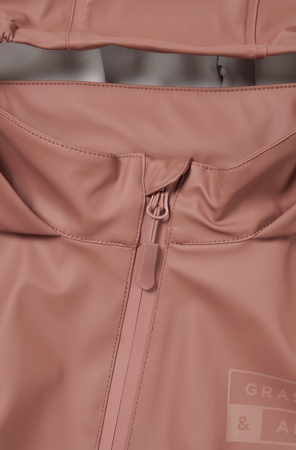 Close-up detail of the back waist panel of the Grass & Air Rose puddle suit; wide horizontal elasticated back waist panel gathered and stitched across the back of the suit creating shape and allowing a generous comfortable fit over clothes and layers; rose waterproof fabric gathered either side of the central elasticated band; taped seam stitching visible; tone-on-tone Grass & Air logo label printed in matching rose visible in the bottom right corner; zip guard flap partially visible at the top of the image