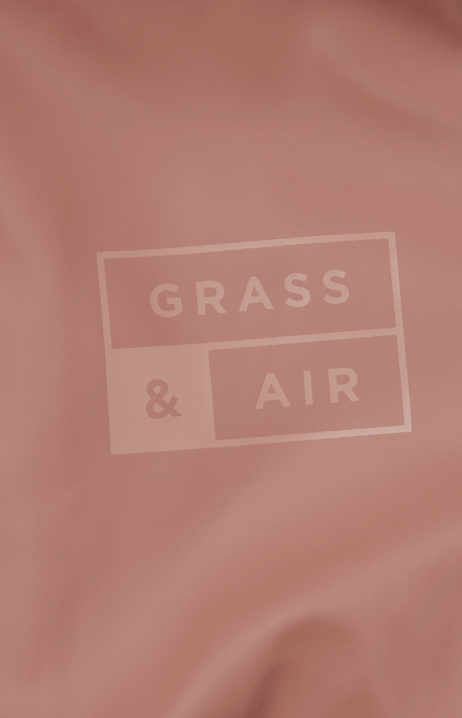 Close-up detail of the tone-on-tone Grass & Air logo printed on the rose puddle suit fabric; the rectangular branded logo features GRASS in the top panel and & AIR in the lower panel in a slightly darker rose on the muted dusty rose waterproof fabric background; the logo is deliberately subtle and understated, printed in matching tones rather than contrasting colour; soft focus rose fabric with gentle folds and creases visible around the central logo; demonstrates the premium branding detail on the Grass & 