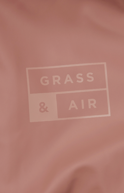 Close-up detail of the tone-on-tone Grass & Air logo printed on the rose puddle suit fabric; the rectangular branded logo features GRASS in the top panel and & AIR in the lower panel in a slightly darker rose on the muted dusty rose waterproof fabric background; the logo is deliberately subtle and understated, printed in matching tones rather than contrasting colour; soft focus rose fabric with gentle folds and creases visible around the central logo; demonstrates the premium branding detail on the Grass & 