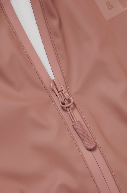Extreme close-up of the waterproof front zip detail on the Grass & Air Rose puddle suit; white silver zip teeth running centrally with a rose-coloured zip pull ring and rose fabric pull tab; a separate rose-coloured waterproof storm zip guard flap running alongside the zip on the right side; taped seam stitching lines visible running parallel to the zip on either side of the opening; tone-on-tone Grass & Air logo text partially visible at the upper right; white hood lining visible at the top of the frame; d