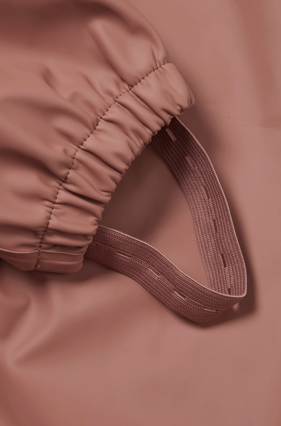 Close-up detail of the integrated boot stirrup and elasticated ankle cuff on the Grass & Air Rose puddle suit; elasticated gathered ankle cuff in rose waterproof fabric ruched around the leg opening at the bottom; wide flat rose-coloured elastic boot stirrup looping downward from the ankle cuff with an open gap at the bottom designed to slip over a welly boot; the stirrup is properly wide and sturdy in matching rose fabric; bokeh soft focus rose fabric background; demonstrates how the boot stirrups keep the