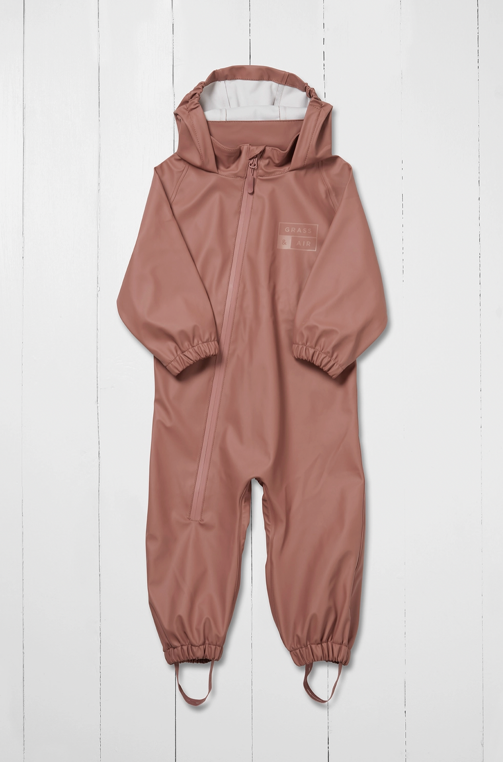 Grass & Air kids waterproof puddle suit in muted dusty rose shown as a front flat lay against a white wooden plank background; full one-piece all-in-one suit in warm clay pink with attached hood folded back and open showing white cream inner lining; waterproof front centre zip running from hood to waist with rose-coloured zip pull; subtle tone-on-tone Grass & Air logo printed on the left chest; long sleeves with elasticated gathered cuffs at the wrists; full legs with elasticated gathered ankle cuffs at the