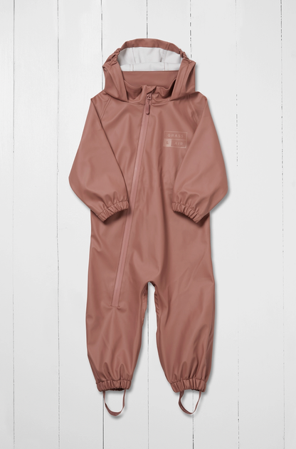 Grass & Air kids waterproof puddle suit in muted dusty rose shown as a front flat lay against a white wooden plank background; full one-piece all-in-one suit in warm clay pink with attached hood folded back and open showing white cream inner lining; waterproof front centre zip running from hood to waist with rose-coloured zip pull; subtle tone-on-tone Grass & Air logo printed on the left chest; long sleeves with elasticated gathered cuffs at the wrists; full legs with elasticated gathered ankle cuffs at the