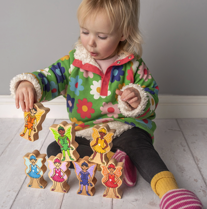Toddler in floral jacket holding orange fairy aloft; 6 remaining fairies on white floor; naturalistic play scene showing scale
