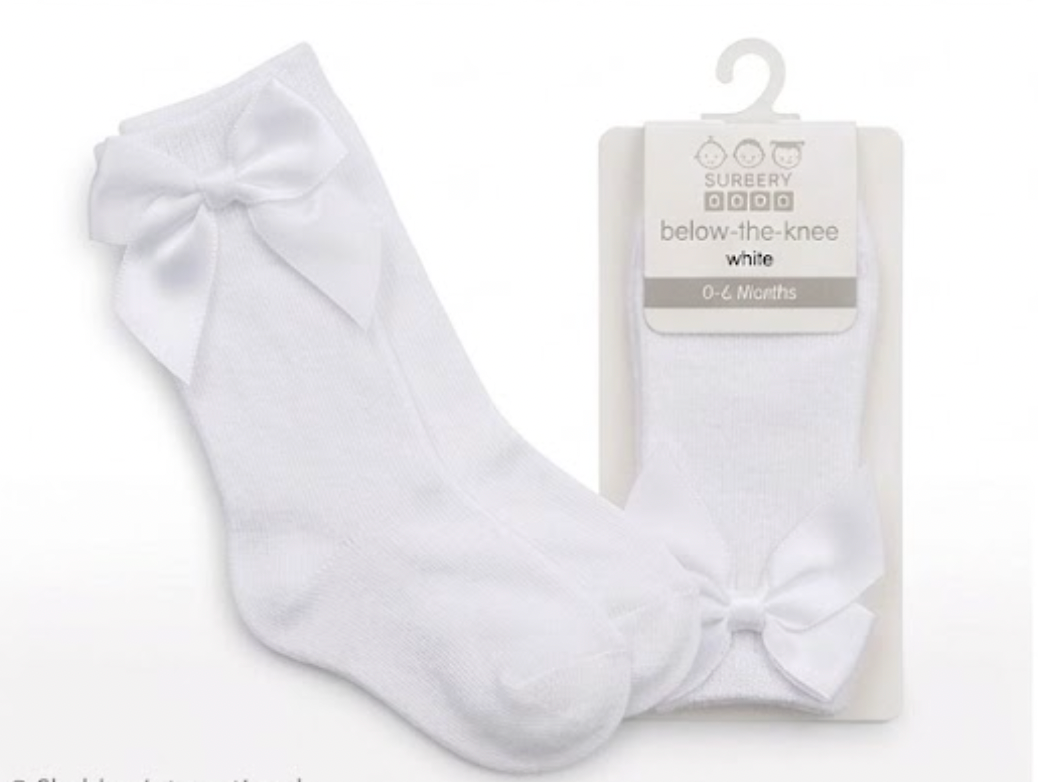 Nursery Time Baby Knee High Socks with Bow in White shown on a white background alongside original retail packaging; crisp white soft fine-knit cotton socks with a long length sitting below the knee; each sock has a large satin ribbon bow in matching white at the back of the calf with two loops and two tails; retail packaging on the right is a white card hanger pack labelled below-the-knee, white, 0 to 6 Months with the Burbery Baby brand mark
