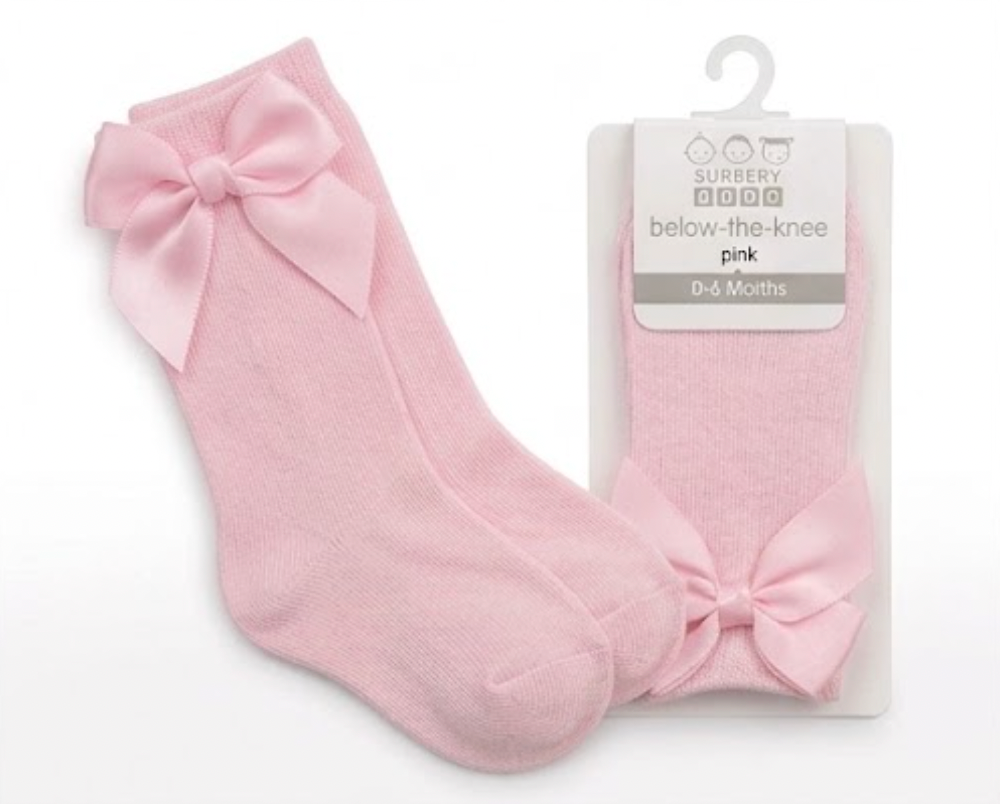 Nursery Time Baby Knee High Socks with Bow in Pink shown on a white background alongside original retail packaging; plain baby-pink soft fine-knit cotton socks with a long length sitting below the knee; each sock has a large satin ribbon bow in matching baby pink at the back of the calf with two loops and two tails; retail packaging on the right is a white card hanger pack labelled below-the-knee, pink, 0 to 6 Months with the Burbery Baby brand mark