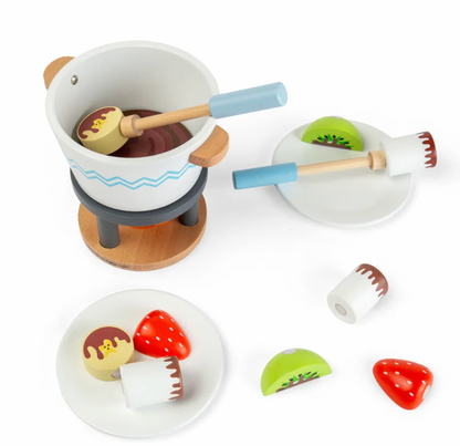 Bigjigs Toys Chocolate Pot for Two on a white background with one dipping stick holding a pale yellow biscuit in the pot; painted orange flame visible on the fire stand below; left plate shows a red strawberry, pale yellow bear-face biscuit and white marshmallow with chocolate drizzle; right plate shows a kiwi slice and a dipping stick; loose strawberry and kiwi pieces to the lower right

