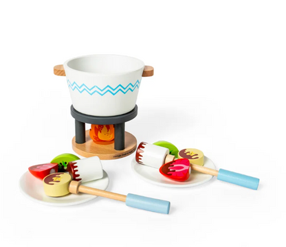 Bigjigs Toys Chocolate Pot for Two 16-piece wooden play fondue set laid out on a white background showing all components; white painted round fondue pot with natural wood ear-shaped handles and a teal chevron band on a dark charcoal three-legged fire stand over a painted orange flame on a round natural wood base; two white round plates holding wooden treat pieces including red strawberry halves, lime green kiwi slices and pale yellow bear-face biscuits with painted chocolate drizzle; two wooden dipping stic