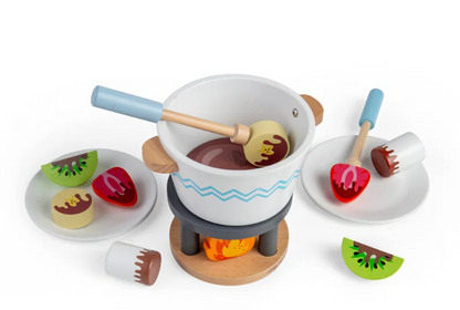 Bigjigs Toys Chocolate Pot for Two showing a dipping stick with sky-blue handle inserted into the white fondue pot, skewering a pale yellow bear-face biscuit piece in the painted chocolate interior; fire stand with painted orange flame visible below; two plates with treat pieces arranged either side; a kiwi slice and white marshmallow with chocolate drizzle in the foreground