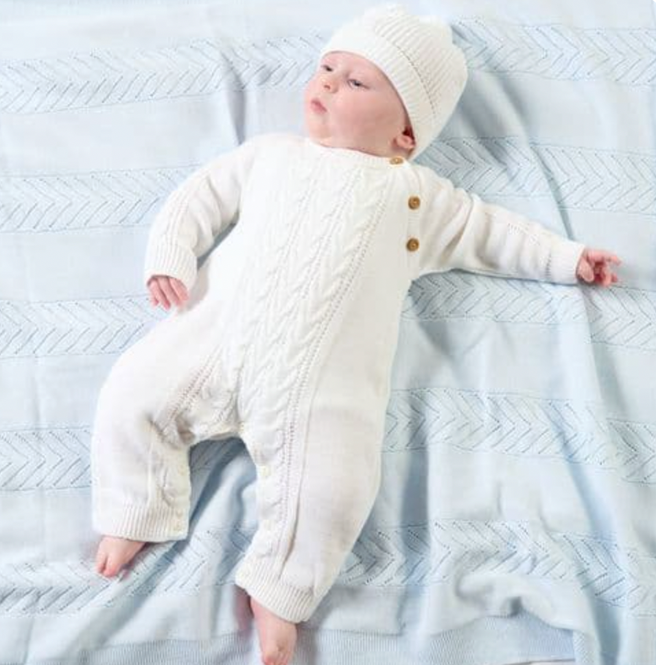Baby lying on a pale blue textured cellular blanket wearing the white cable-knit onesie and matching pompom beanie; arms spread wide; central cable panel, three wooden shoulder buttons and ribbed ankle cuffs all clearly visible; bare feet below the cuffs