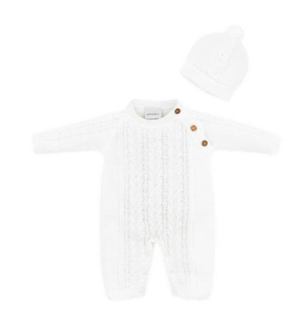 Dandelion Knitted Cable Knit Baby Onesie and Hat Set in White shown as a flat lay on a white background; full-length onesie in bright white acrylic knit with a central cable-knit panel running from chest to ankle, flanked by smoother knit side panels; three round wooden buttons at the right shoulder; Dandelion woven label at the neckline; ribbed cuffs at wrists and ankles; matching white close-fitting beanie with a small pompom at the crown
