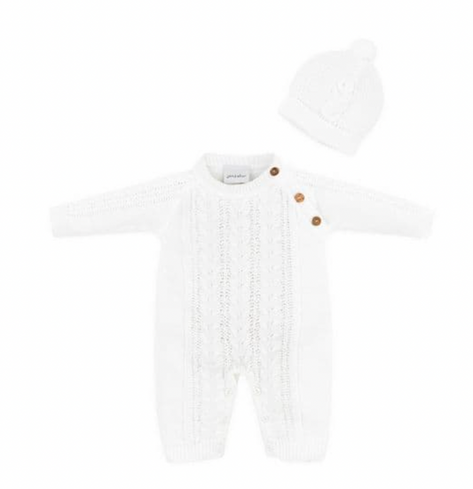 Dandelion Knitted Cable Knit Baby Onesie and Hat Set in White shown as a flat lay on a white background; full-length onesie in bright white acrylic knit with a central cable-knit panel running from chest to ankle, flanked by smoother knit side panels; three round wooden buttons at the right shoulder; Dandelion woven label at the neckline; ribbed cuffs at wrists and ankles; matching white close-fitting beanie with a small pompom at the crown
