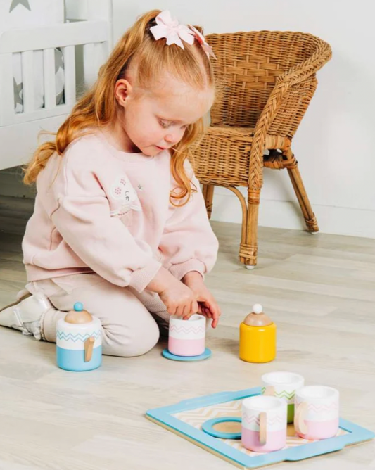 Child playing with the Bigjigs wooden tea set on the floor, placing a wooden spoon in a pink cup beside the blue teapot, yellow sugar pot and serving tray.