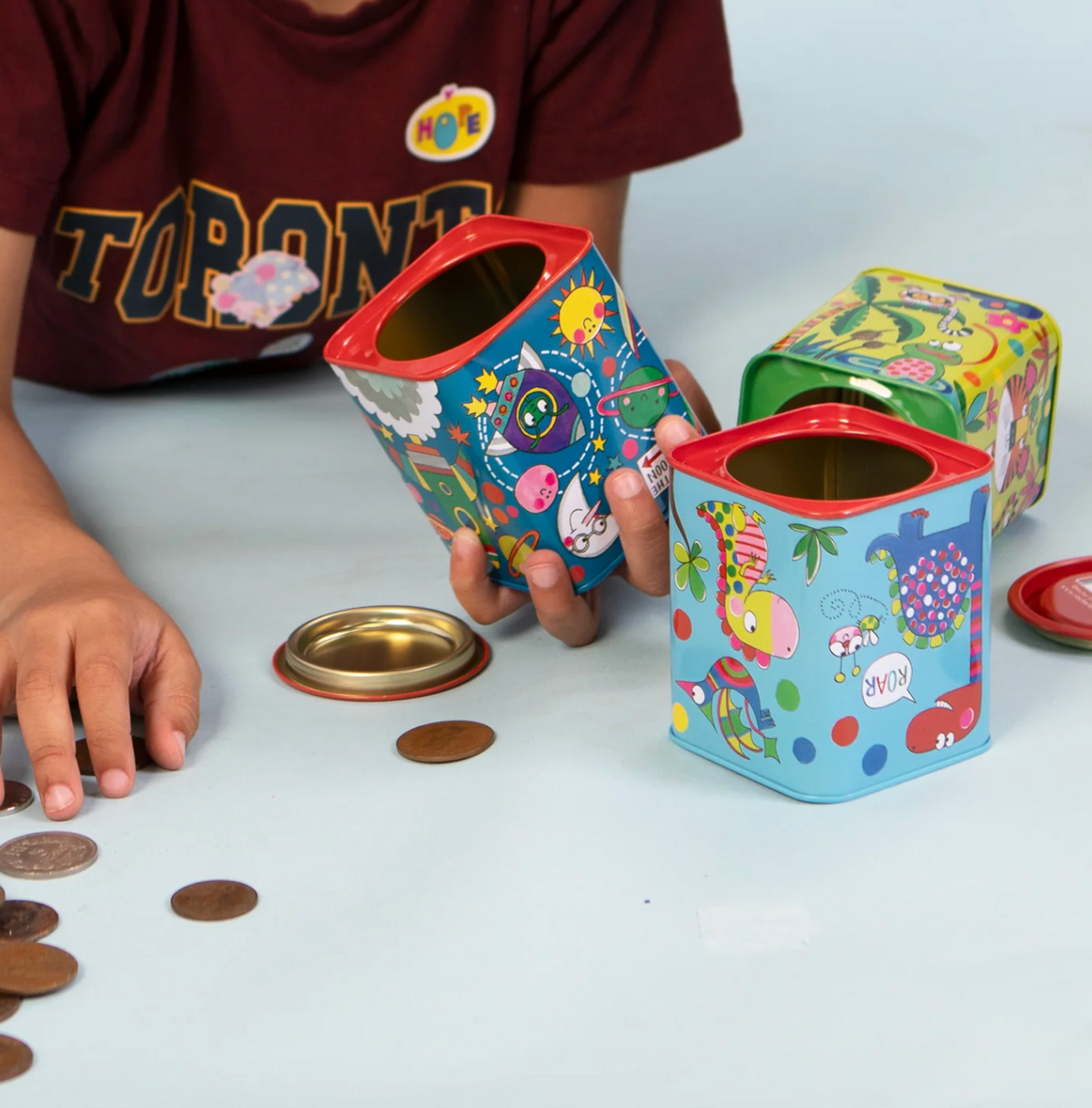 Colourful Rachel Ellen money box tins arranged together, including a blue dinosaur tin with playful animal illustrations and a coin slot