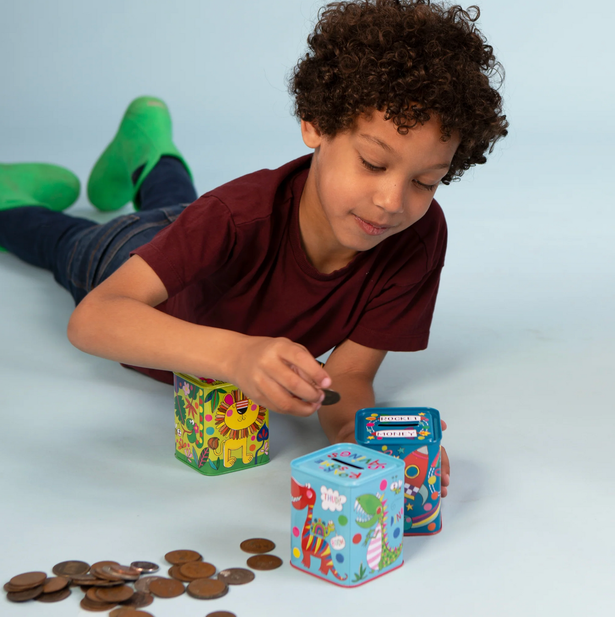 Child using a colourful Rachel Ellen dinosaur money box tin alongside other illustrated money boxes, with coins and a removable base on the table