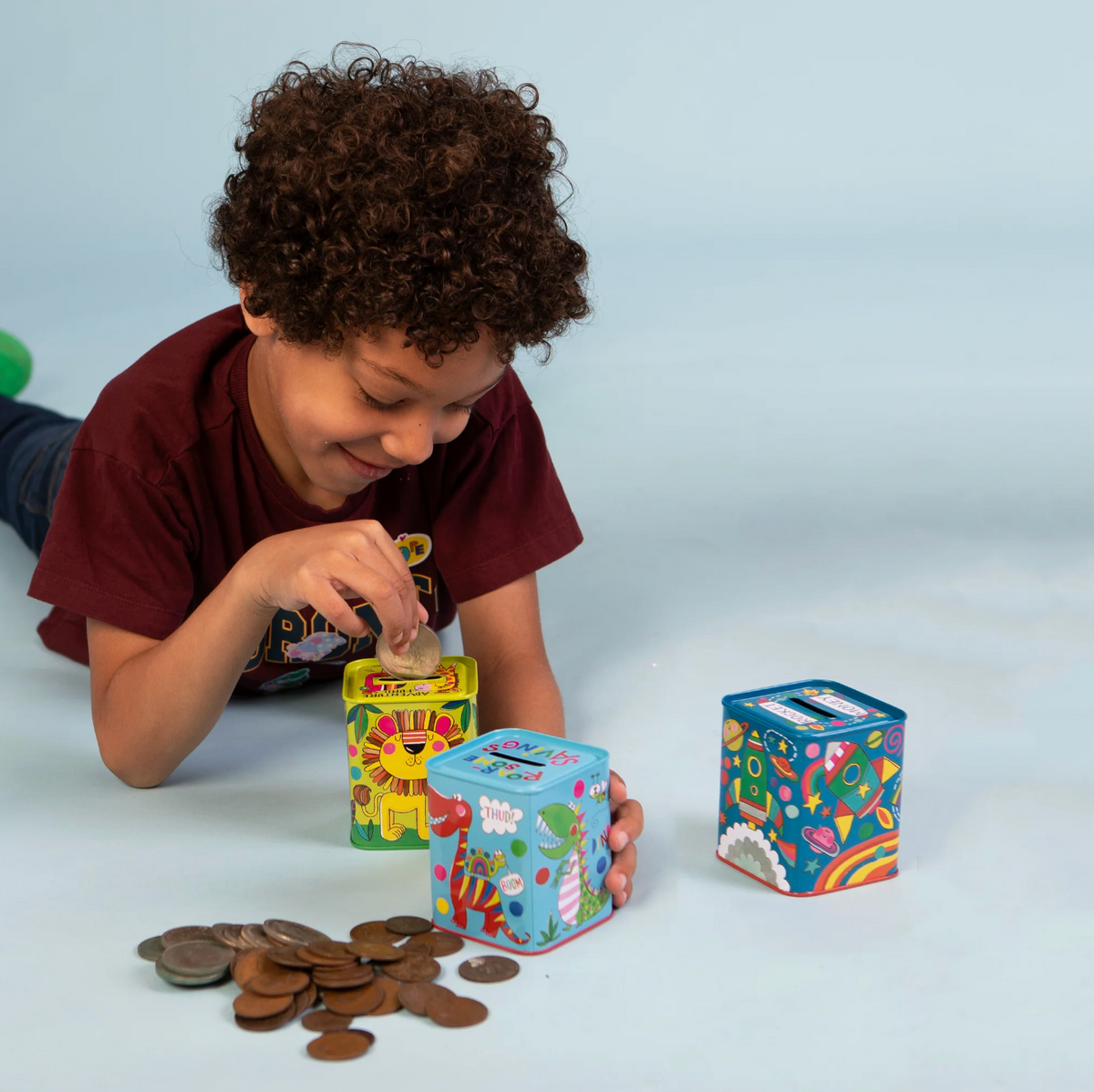 Rachel Ellen money box collection showing the blue dinosaur tin, rocket design and other colourful illustrated savings tins with removable bases