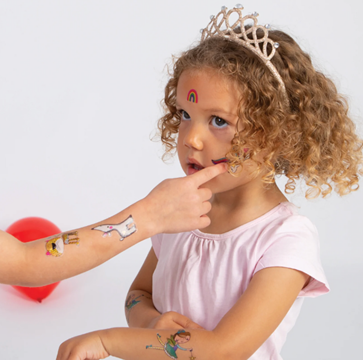 Young child wearing a tiara and smiling while displaying Rachel Ellen Love Our Planet temporary tattoos, including rainbow, leopard, white animal and colourful designs on the arms and face