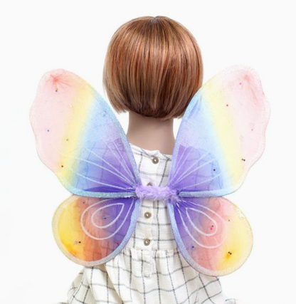 Child wearing Molly & Rose pastel rainbow fairy wings over a checked dress; wings blend pink, peach, yellow, blue and lilac tones with frosty glitter and sequin stars
