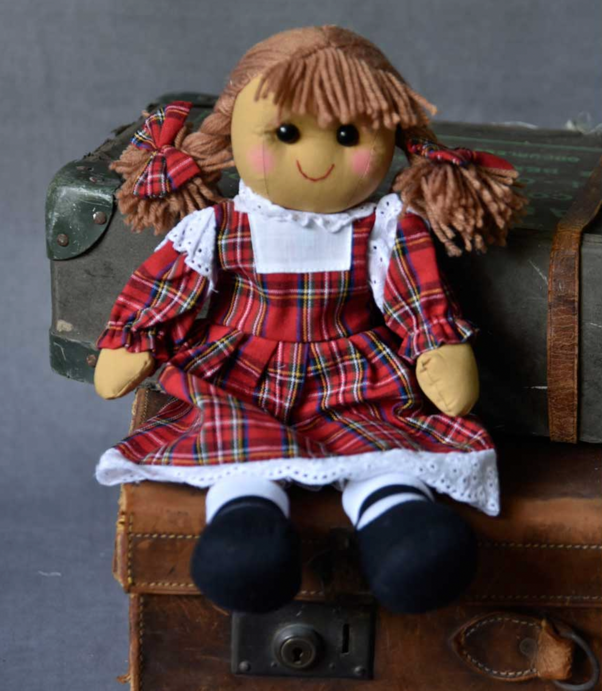 Powell Craft tartan dress rag doll seated in a lifestyle setting, wearing a red tartan dress with white broderie anglaise lace trim, black shoes and tartan hair bows.
