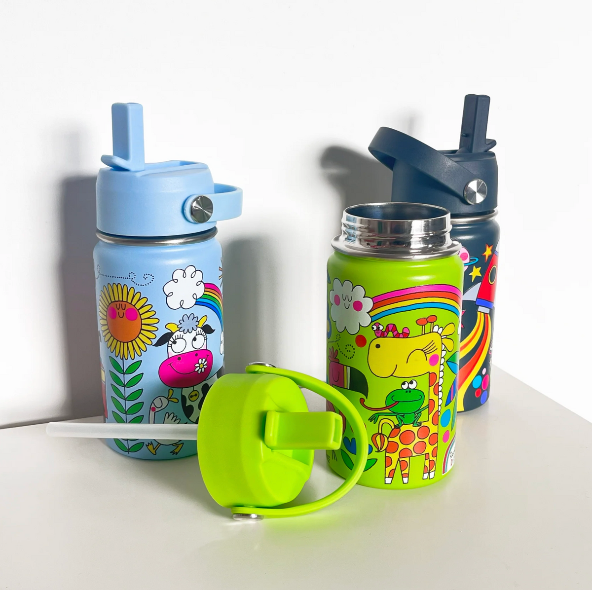 Rachel Ellen bottle range on a white shelf: sky-blue On the Farm bottle at left, open lime-green giraffe-and-frog bottle in front and navy space bottle behind; detached green lid and long clear straw lie in the foreground.