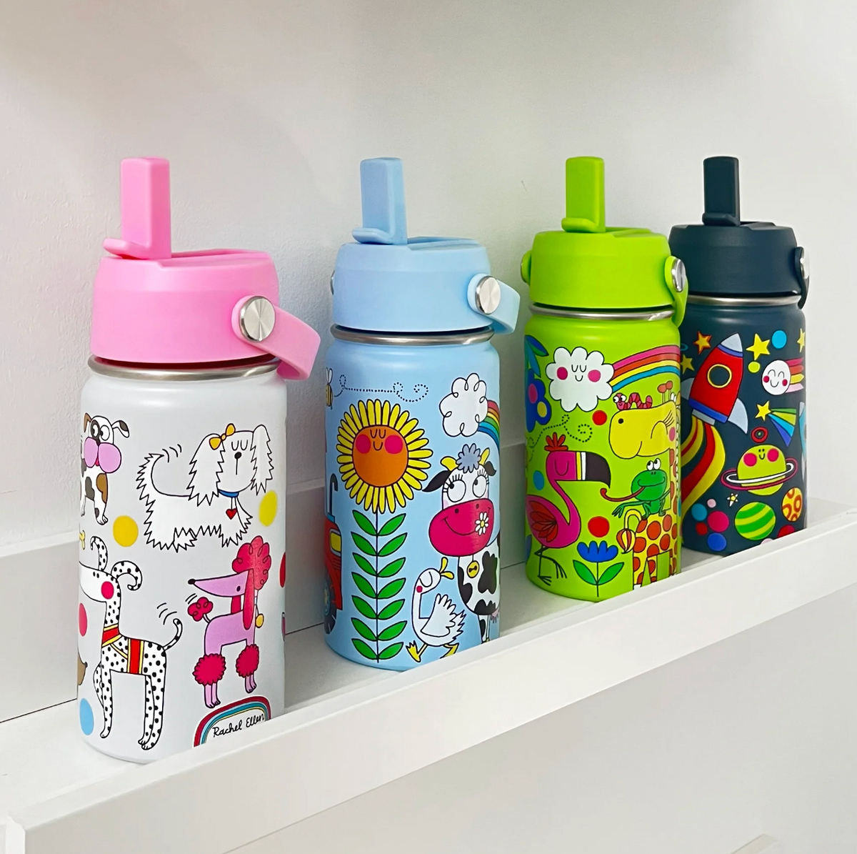 Four Rachel Ellen water bottles on a white shelf, left to right: white dog-print bottle with pink lid, sky-blue On the Farm bottle with sunflower and cow, lime-green animal bottle and navy space bottle; all straws raised.