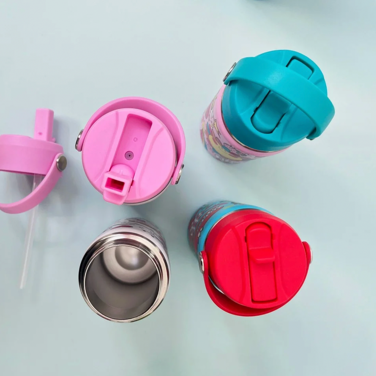 Overhead bottle detail on pale blue surface: detached pink straw lid and clear drinking tube at left, pink raised-straw lid above an open stainless steel bottle, turquoise closed lid at upper right and red closed lid below.