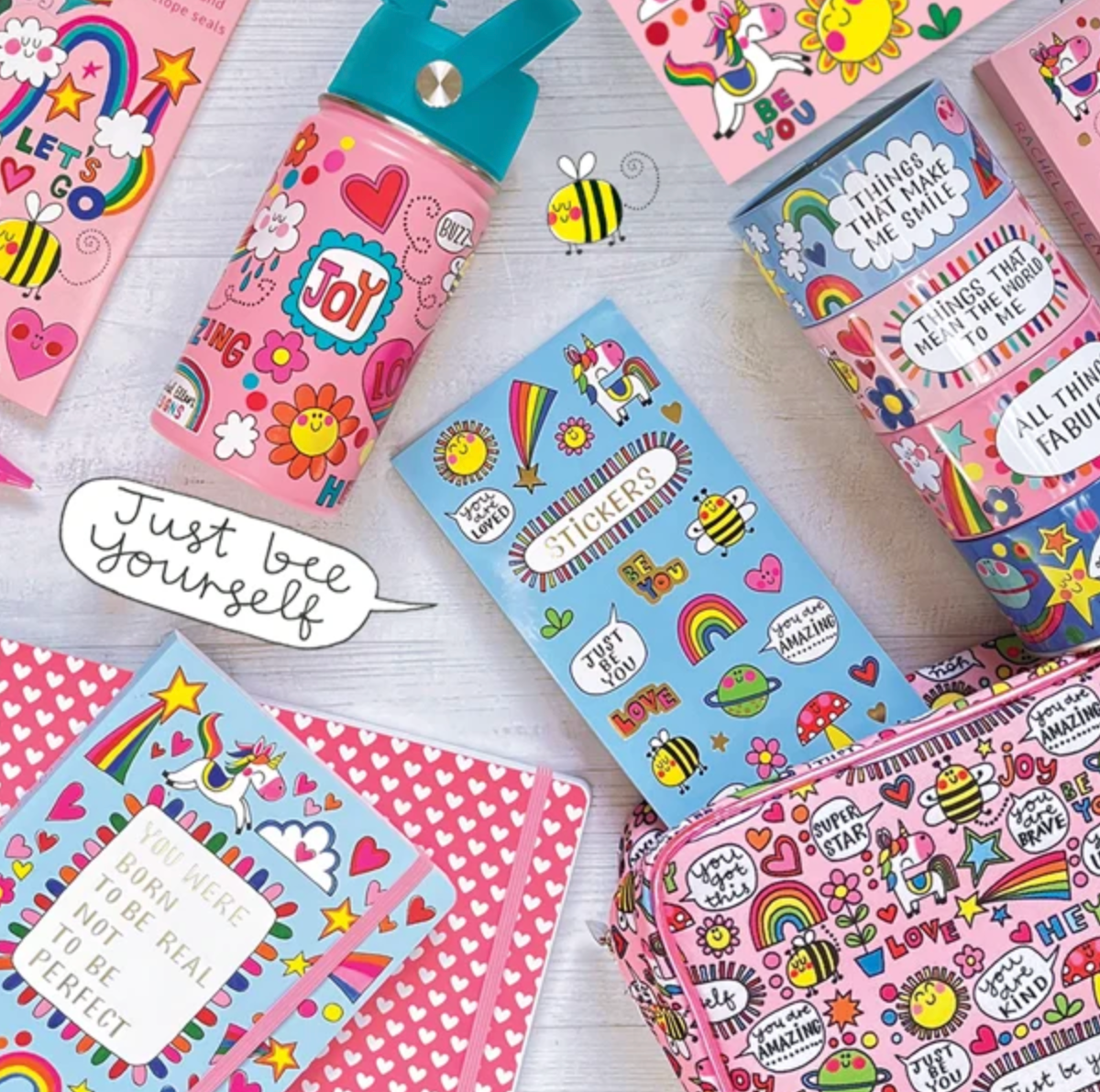 Rachel Ellen Bee Happy pink bottle with turquoise lid at upper left of a colourful stationery flat lay; JOY and BUZZ visible on bottle, blue STICKERS sheet in centre, stacked keepsake boxes at right and pink zipped case below.