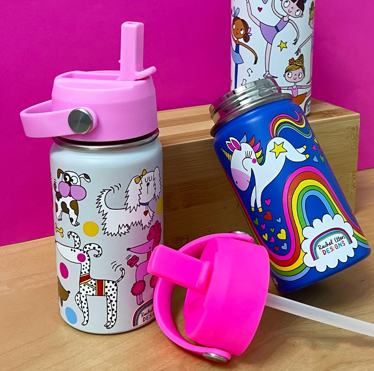 Rachel Ellen bottles on a wooden display against a bright pink backdrop: open cobalt-blue Unicorn bottle leaning at right, white dog-print bottle with pink lid at left and ballet bottle behind; detached fuchsia lid and clear straw in front.