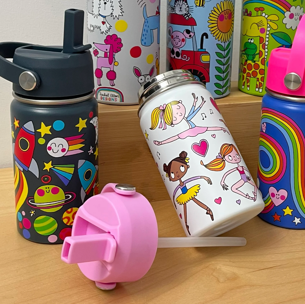 Open Rachel Ellen Ballet white bottle leaning on a wooden display, illustrated with dancers and pink hearts; detached pink straw lid and clear tube in foreground, navy space bottle at left, blue rainbow bottle at right and dog, farm and animal bottles behind.⠋